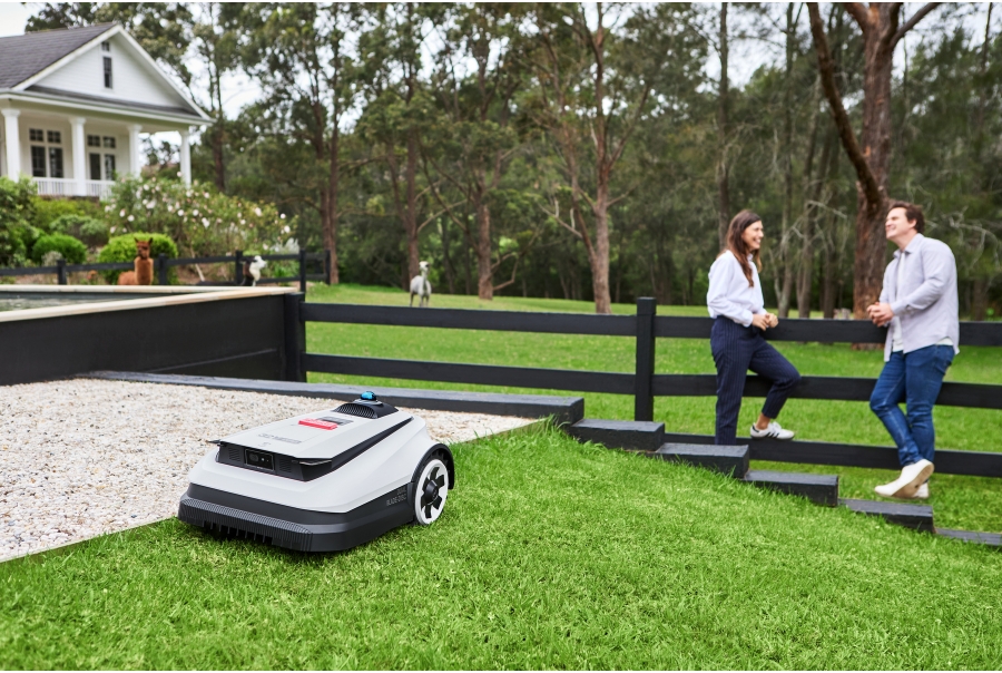 Un robot tondeuse à gazon en forme de chèvre tond une pente devant un couple